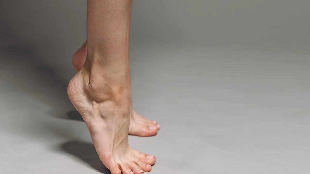 Side view of a bare lower leg and foot raised onto the toes with the heel lifted, against a plain grey background